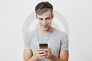 Handsome caucasian young male with dark hair and blue eyes holding mobile phone, smiling with teeth, typing message to