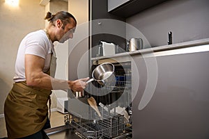 Handsome Caucasian young man in beige chef apron, takes out a clean saucepan from an overload automatic dishwasher with