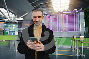 Handsome Caucasian young man, business traveler using smartphone, booking hotel, standing with his back to flight information