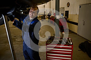 Handsome Caucasian young man, car engineer, technician, auto mechanic repairing a lifted modern automobile on a hoist in the