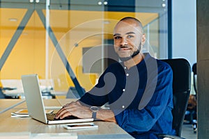 Handsome cheerful african american man in creative atmosphere using laptop sitting on a wooden table