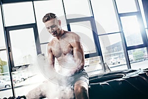 handsome cheerful shirtless sportsman sitting on tire and applying talcum powder on hands