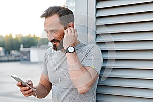 Handsome cheerful strong mature sportsman listening music with earphones using mobile phone.
