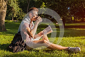 Handsome college man reading a book and drinking coffee in campus park. Happy guy student learning sitting on grass.
