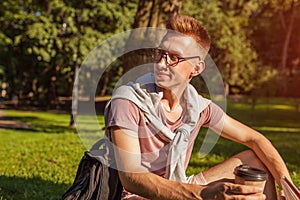 Handsome college man reading a book and drinking coffee in campus park. Happy guy student learning sitting on grass.