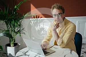 Handsome computer programmer sitting at workplace. Successful freelancer copywriter working from home. Pensive man wearing eyeglas