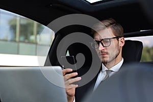 Handsome confident man in full suit looking at his smart phone while sitting in the car and using laptop