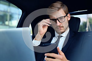 Handsome confident man in full suit looking at his smart phone while sitting in the car and using laptop.