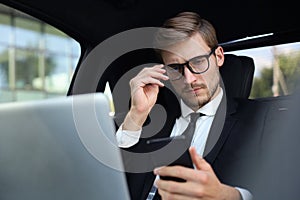 Handsome confident man in full suit looking at his smart phone while sitting in the car and using laptop