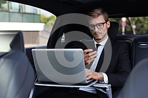 Handsome confident man in full suit looking at his smart phone while sitting in the car and using laptop