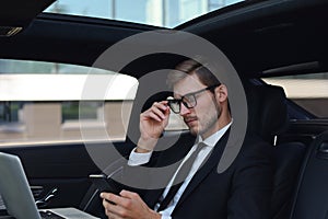 Handsome confident man in full suit looking at his smart phone while sitting in the car and using laptop