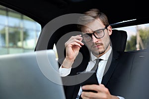 Handsome confident man in full suit looking at his smart phone while sitting in the car and using laptop