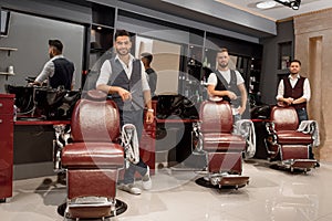 Confident masters barbers standing near hairdresser chairs and posing.