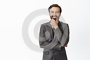 Handsome corporate man in suit looking pleased, smiling and touching his beard with satisfied face expression, likes
