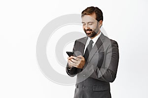 Handsome corporate man in suit using mobile phone, chatting or ordering in application, standing over white background