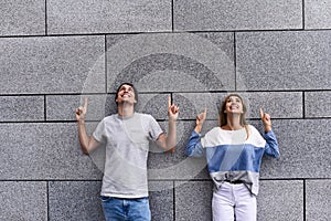 Handsome couple is pointing away and smiling, standing against gray wall.