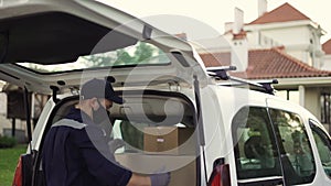 Handsome courier putting cardboard box package in delivery van and enroll the parcel. Courier in mask, cap and gloves on