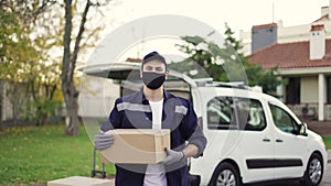 Handsome courier takes cardboard box package out of delivery van walks through private sector. Courier in mask, cap and