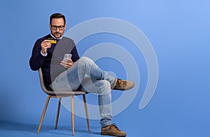 Handsome customer making online payment with cellphone and credit card on chair over blue background