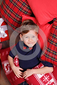 Handsome cute boy celebrating New Year Christmas alone close to xmas tree on red pillow posing in studio decoration wearing jeans