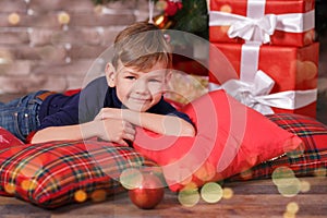 Handsome cute boy celebrating New Year Christmas alone close to xmas tree on red pillow posing in studio decoration wearing jeans