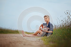 Handsome cute boy is playing on acoustic guitar sittingon road in summer day.