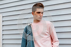 Handsome cute young man model in trendy pink sweatshirt in fashion denim blue youth jacket posing near white wall outdoors. Urban