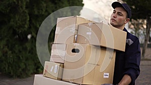 Handsome delivery man in uniform, cap and gloves carrying many heavy cardboard boxes parcels outdoor