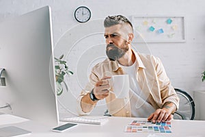 Handsome designer drinking beverage and looking at computer monitor