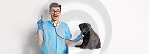 Handsome doctor veterinarian smiling, examining pet in vet clinic, checking pug dog with stethoscope, pointing finger up