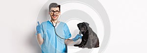 Handsome doctor veterinarian smiling, examining pet in vet clinic, checking pug dog with stethoscope, showing thumbs-up