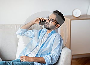 Handsome Eastern Man Talking On Cellphone While Relaxing On Sofa At Home