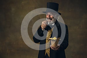 Handsome elderly gray-haired man, gentleman, aristocrat or actor eating fast food isolated on dark vintage background