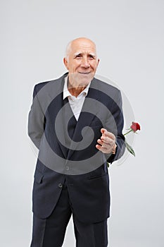 A elderly man in a black suit, smiling and holds one red rose behind him. Isolation.