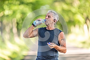 Handsome elderly man rehydrates after a run outdoors in the nature by drinking water from a bottle