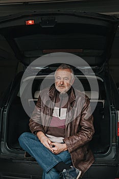 Handsome elderly man sitting in open trunk of car