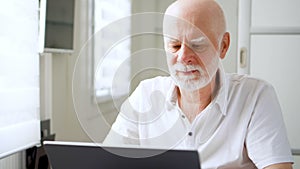 Handsome elderly senior man working on laptop computer at home. Remote freelance work on retirement