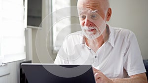 Handsome elderly senior man working on laptop computer at home. Remote freelance work on retirement