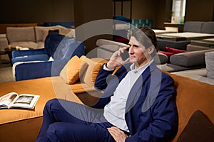 Handsome elegant imposing mature man in a business suit talking on the phone while sitting on a sofa in a furniture store