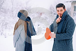 Handsome elegant man, finger to mouth - tsss gesture, surprise for his girlfriend holding red heart, greeting card with