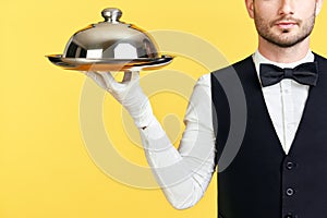Handsome elegant waiter holding tray and cloche ready to serve on yellow background