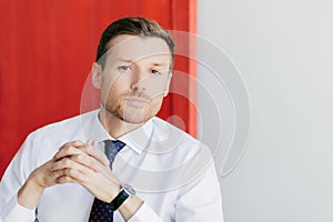 Handsome employee with stubble contemplates business project, holds hands together. Poses against red and white background. Copy