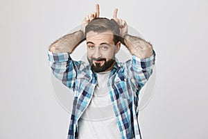Handsome european adult guy with beard and moustache showing horns with index fingers on hand, imitating devil or goat