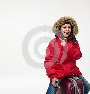 Handsome European boy dressed in bright red parka holding airplane toy model and pointing on a copy space of white background