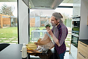 Handsome European man holding greens while unpacking eco paper bag with healthy food, standing at kitchen countertop in a spacious