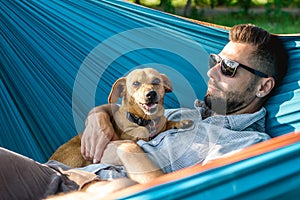 Handsome European man in sunglasses is resting in hammock with his cute little dog