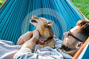 Handsome European man in sunglasses is resting in hammock with his cute little dog