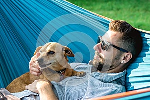 Handsome European man in sunglasses is resting in hammock with his cute little dog