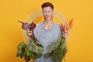 Handsome fairhaired man holding fresh vegetables in hands, young raw foodist decides what to eat beets or carrots isolated over photo