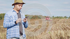 Handsome farmer with smartphone standing in field sunflower with combine harvester in background. Concept modern
